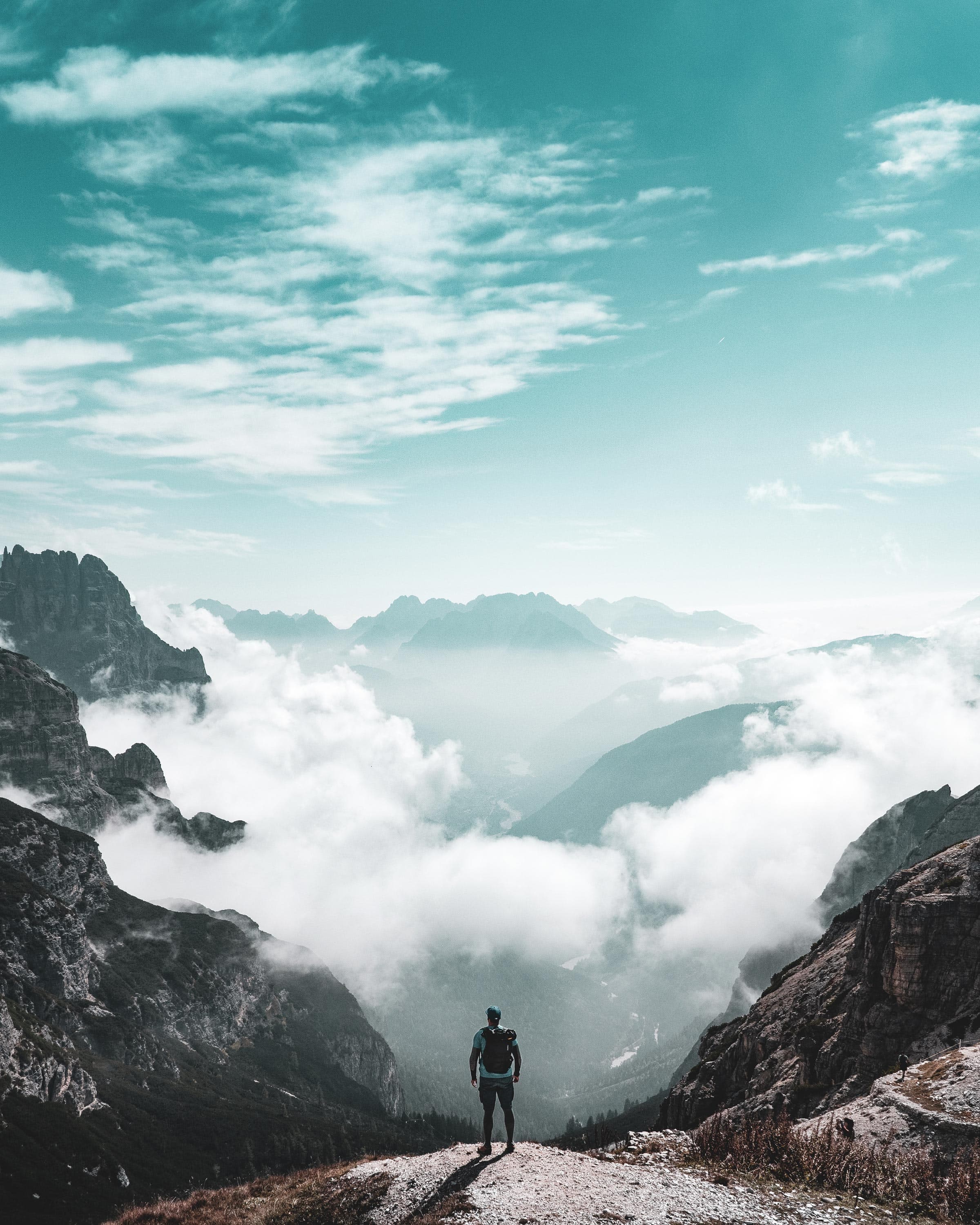 A person stands at the edge of a cliff, looking out over a deep valley filled with clouds and surrounded by tall, jagged mountains under a blue sky with scattered clouds.
